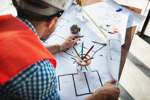 Man Wearing White Hard Hat Leaning on Table With Sketch Plans