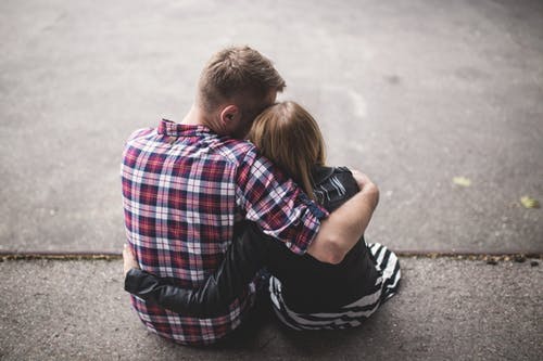 Man in Red White and Blue Check Long Sleeve Shirt Beside Woman in Black and White Stripes Shirt Hugging Each Other While Sitting on a Concrete Surface