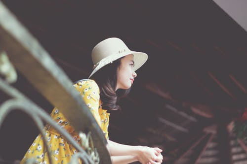 Photo of Woman in Sun Hat and Floral Dress Leaning on Metal Railing Looking Out
