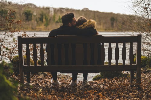 Man Kissing Woman's Head While Sitting on Bench in Front of Body of Water